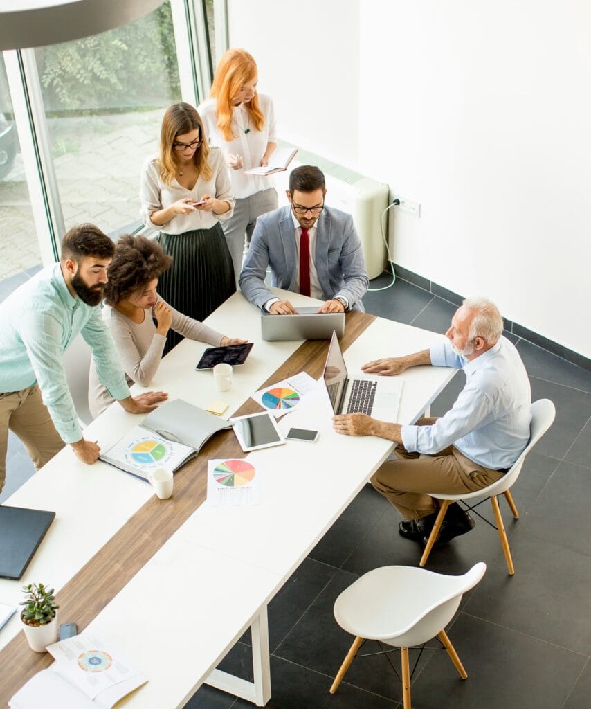 Équipe de sept personnes réunie autour d'une longue table de travail avec portables et graphiques imprimés