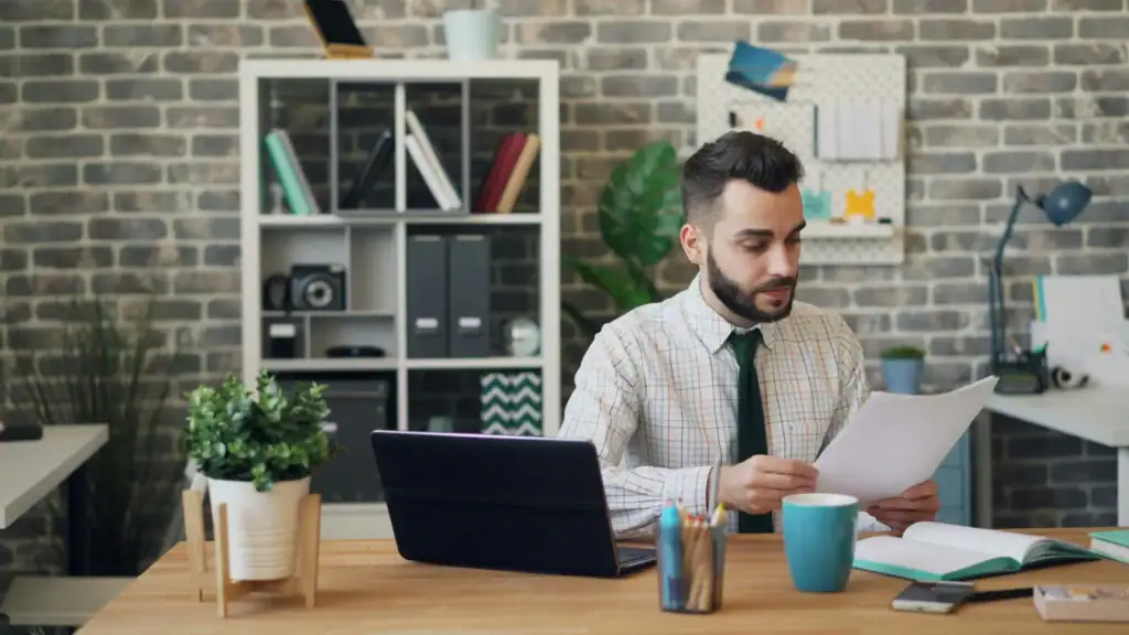 Dirigeant de PME qui compare des documents d'assurance à son bureau, ordinateur portable ouvert devant lui